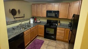 Cocina compacta con encimeras de granito, gabinetes de madera, estufa negra, microondas y alfombra morada.