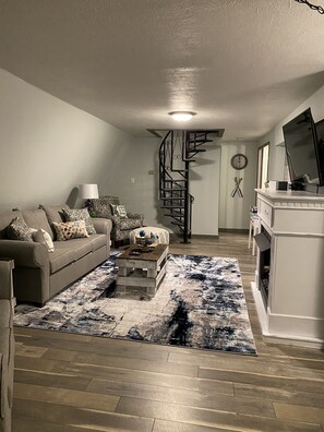 Ein Wohnzimmer mit Sofa, Couchtisch und einem gemusterten Teppich. Eine Wendeltreppe führt zu einer oberen Ebene.