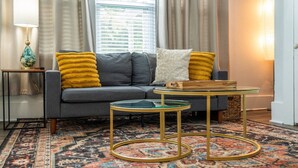 A living room with a grey sofa, yellow throw pillows, a glass coffee table, a patterned rug, and a lamp on a side table. - Cedar Blossom Bungalow - Pet friendly getaway with a 5 min walk to downtown (Hot Springs)