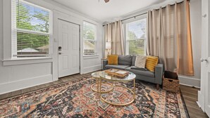A living room with a gray sofa, a patterned rug, a glass coffee table, and a ceiling fan. - Cedar Blossom Bungalow - Pet friendly getaway with a 5 min walk to downtown (Hot Springs)