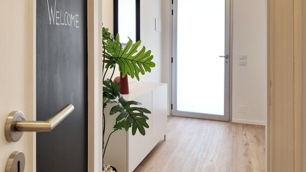 Un couloir avec une pancarte « BIENVENUE », une plante en pot, un miroir et une porte qui donne sur une pièce lumineuse.