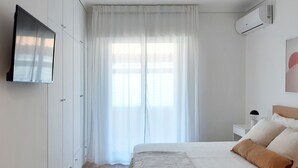 Une chambre à coucher comprenant un lit, un téléviseur fixé au mur, une fenêtre avec des rideaux légers et un appareil de climatisation.