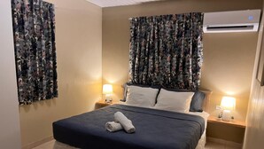 A hotel room with a bed, two bedside tables with lamps, floral curtains, and an air conditioning unit on the wall. - Aruba Sun, Sand & Beach  Apartment 5 min from Beach. (Sanicolas)