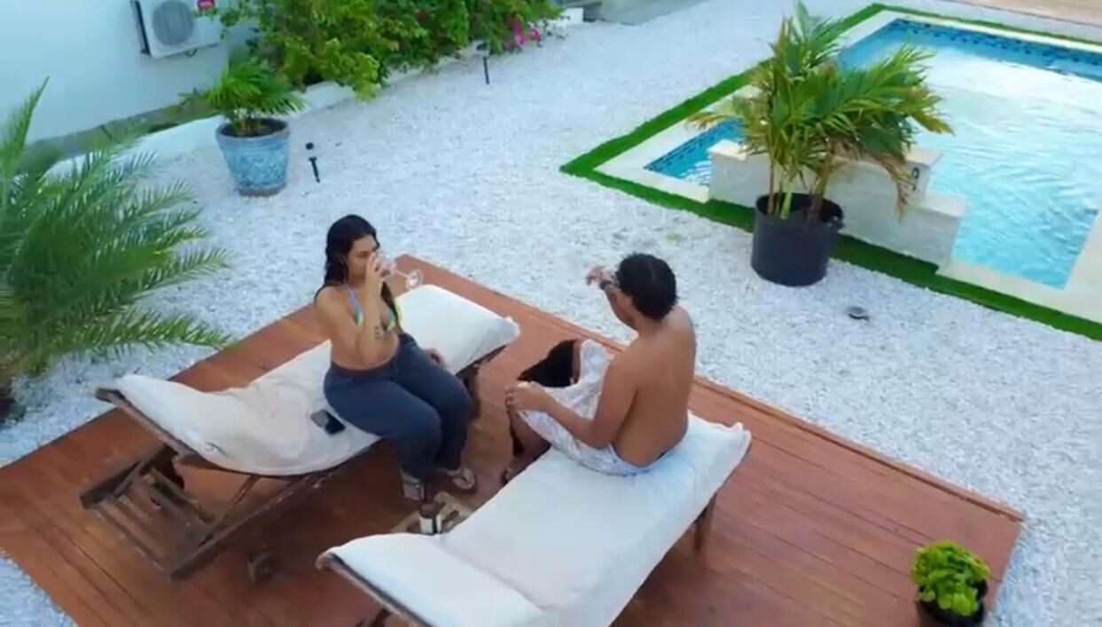Dos personas sentadas en sillones junto a una piscina, con un pequeño jardín y una piscina al fondo.