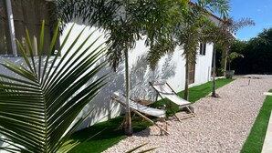 A patio area with two lounge chairs, a palm tree, and a white building with a window. - Aruba Sun, Sand & Beach  Apartment 5 min from Beach. (Sanicolas)