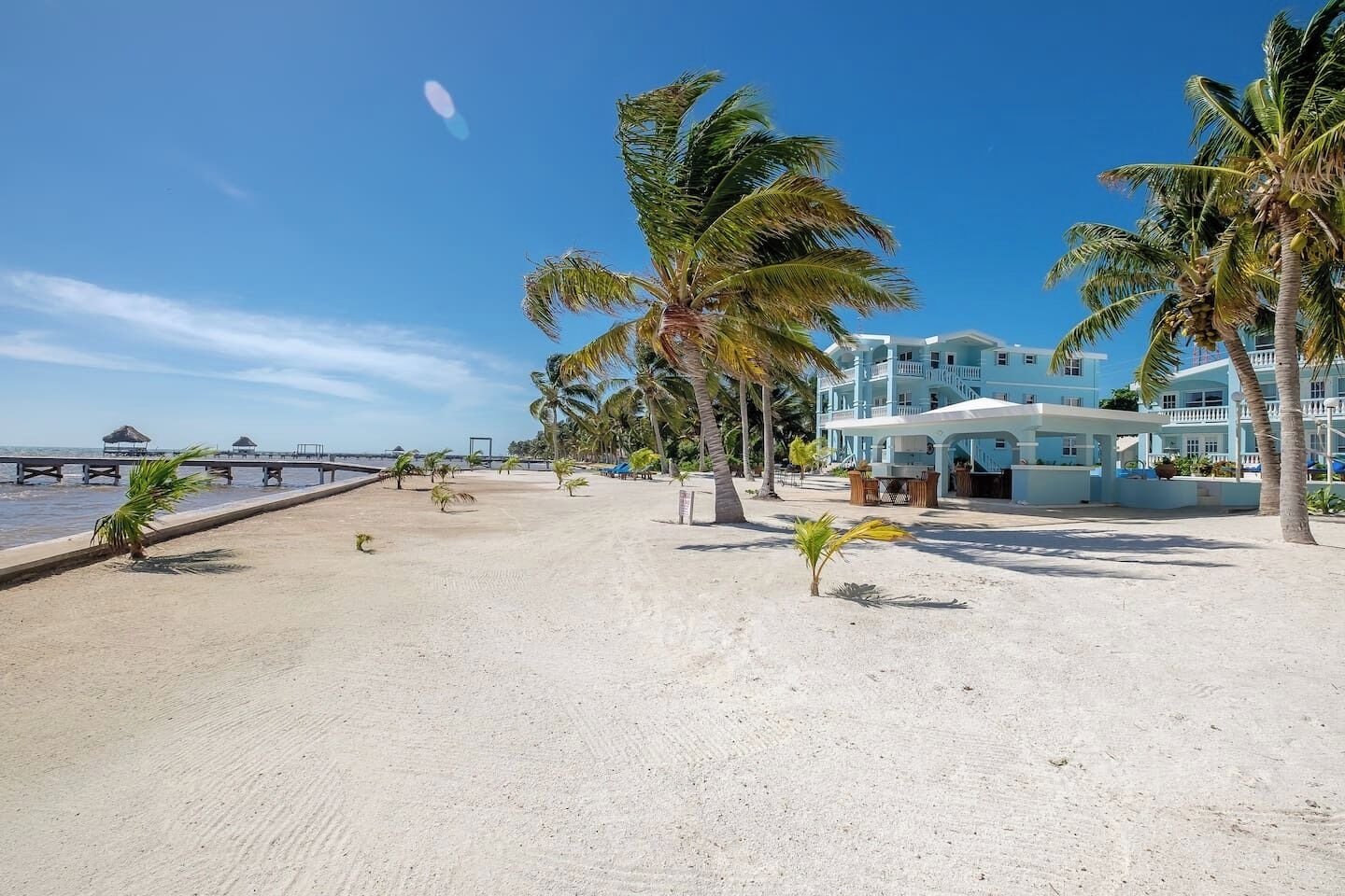 A beachfront hotel with a sandy beach, palm trees, and a pier in the background.