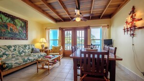 A living room with a sofa, coffee table, and dining table with chairs. There is a ceiling fan, a painting, and a TV on the wall. - Charming 3 Bed Condo with Exquisite Caribbean View (San Pedro Town)