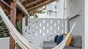 Una cadira de fil a l'ombra d'una pérgola de fusta, un sofà gris amb coixins i un paviment de rajoles amb dibuixos.