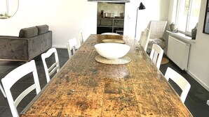Un comedor con mesa de madera rústica, sillas blancas, lámpara colgante y cocina al fondo.