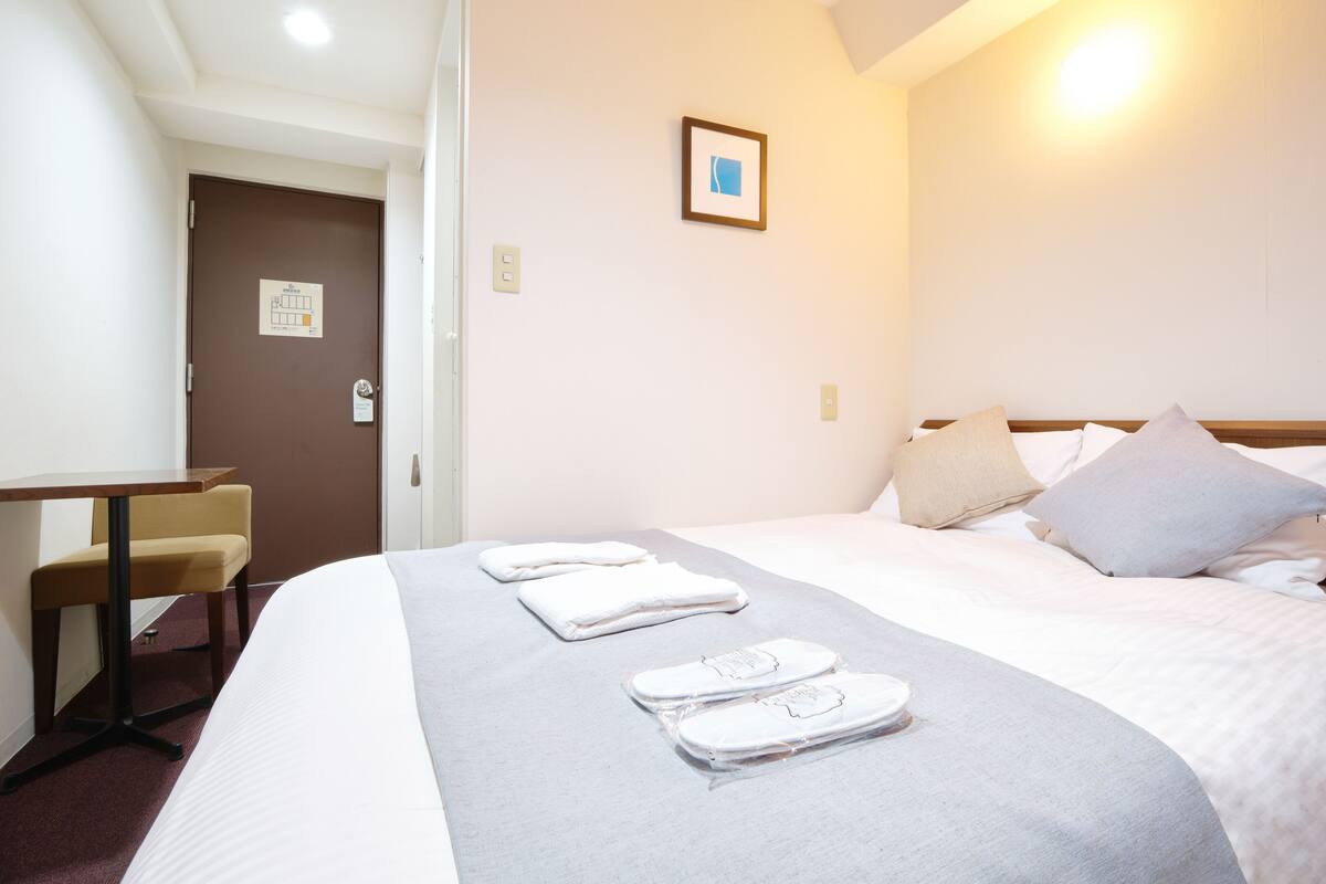 A hotel room with a bed, a desk, and a chair. There is a framed picture on the wall and a door with a sign.