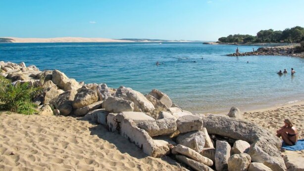 Une plage avec de gros rochers au premier plan, une étendue de sable et des personnes qui se baignent dans l’eau.