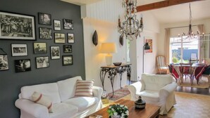 Un salon avec un canapé blanc, une table basse en bois et un lustre. Des tableaux encadrés ornent le mur, et on aperçoit un coin repas au fond de la pièce.