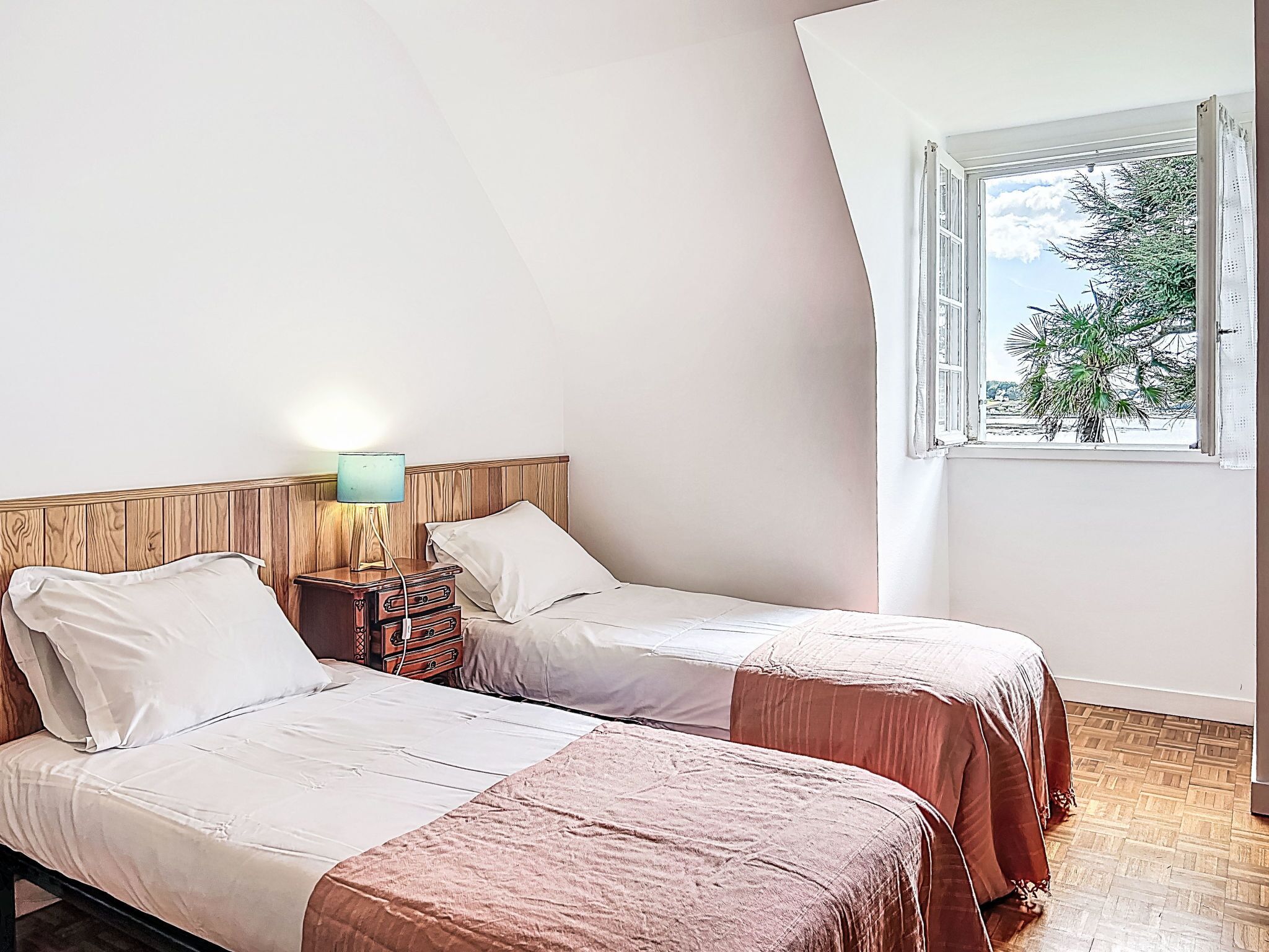 Un dormitorio con dos camas, una mesita de noche con lámpara, una ventana con vistas a árboles, y suelo de madera.