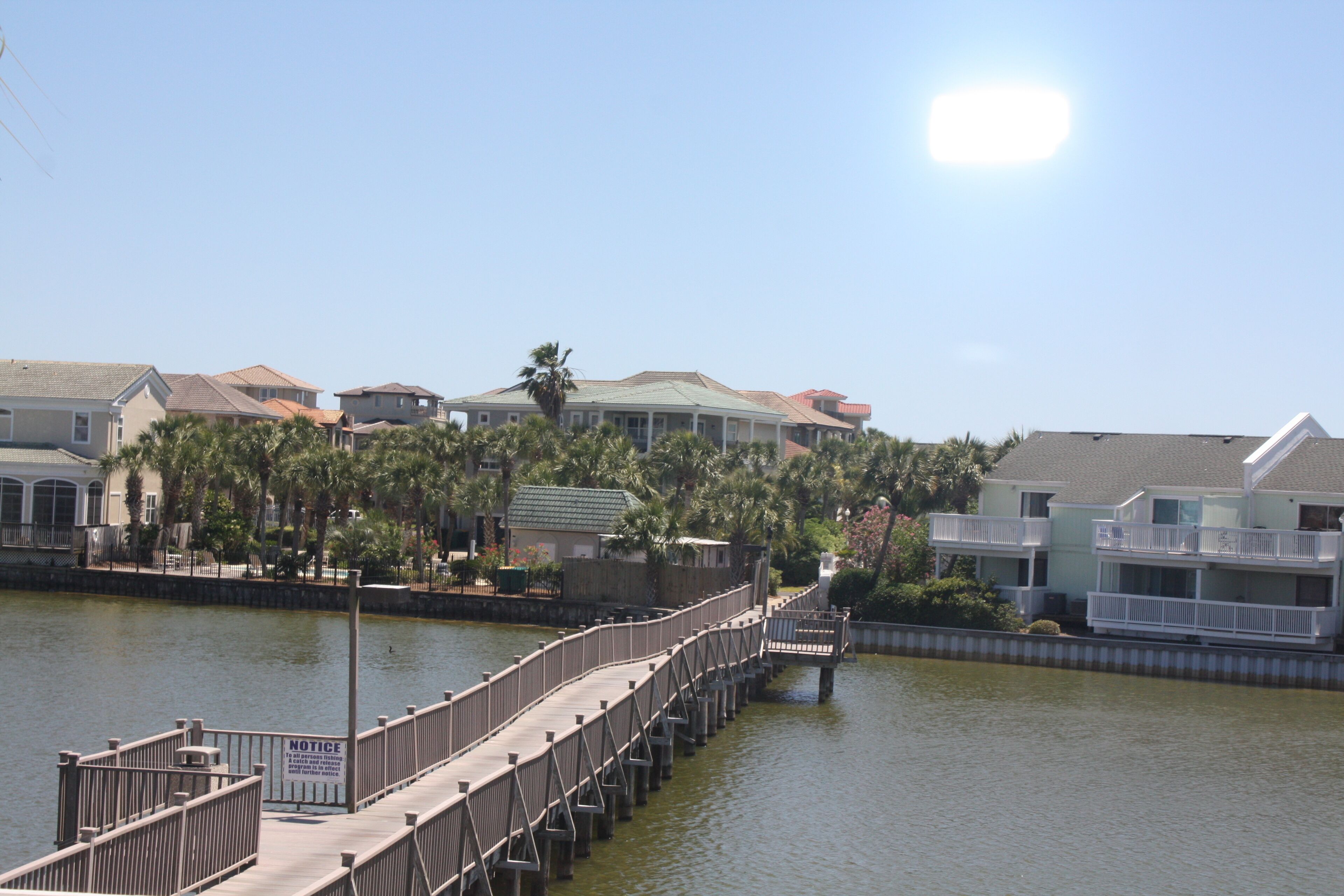 A waterfront property with a wooden pier leading to a dock, surrounded by residential buildings and palm trees.