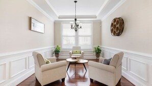 A room with two beige armchairs, a small round table, a chandelier, and a window with blinds.