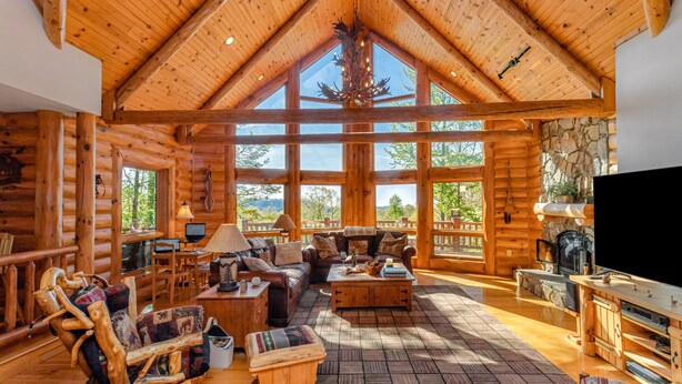A log cabin interior with a large living area, wooden furniture, a stone fireplace, and large windows offering a view of the outdoors.