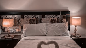 Habitación de hotel con cama, dos lámparas de noche, un cuadro en la pared y una decoración con toallas en forma de corazón sobre la cama.