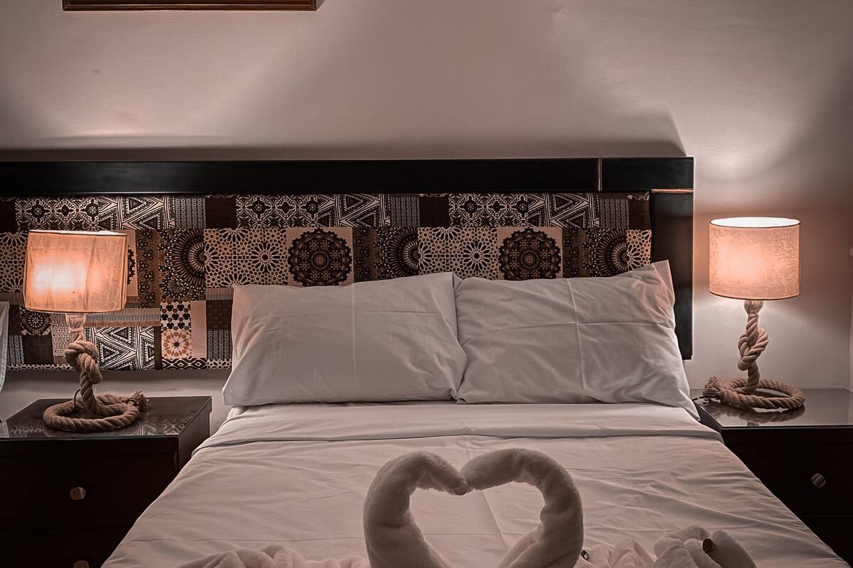 A hotel room with a bed, two bedside lamps, a framed picture on the wall, and a heart-shaped towel arrangement on the bed.