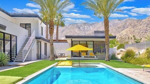 Una casa moderna con piscina, palmeras y montañas al fondo.