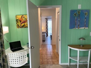A hallway with a laptop on a white stand, a door leading to another room, and a painting of palm trees on the wall. - Three Palms (Kailua-Kona)