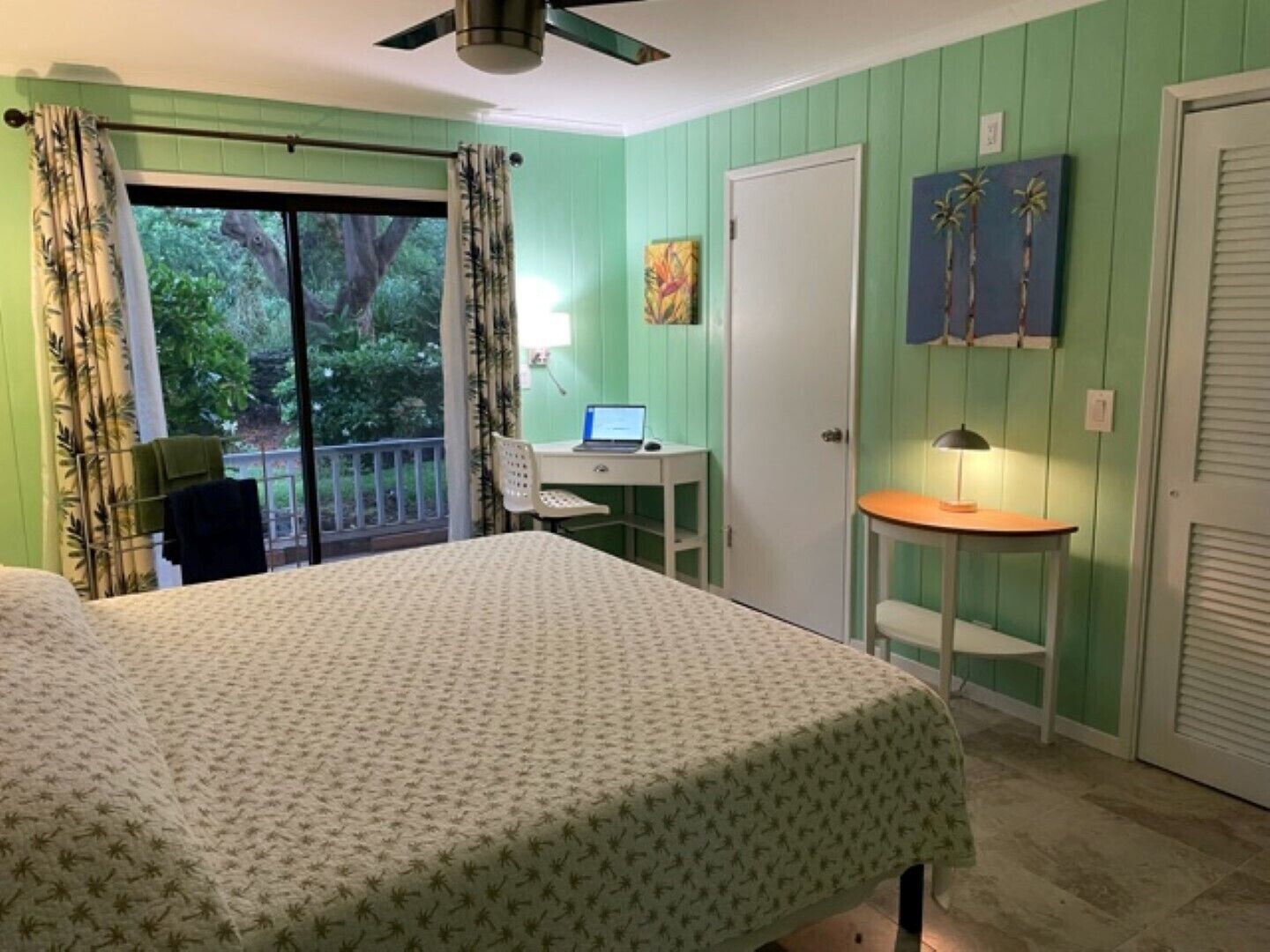 Une chambre à coucher avec un lit, un bureau avec un ordinateur portable, une chaise, une petite table avec une lampe et une porte.