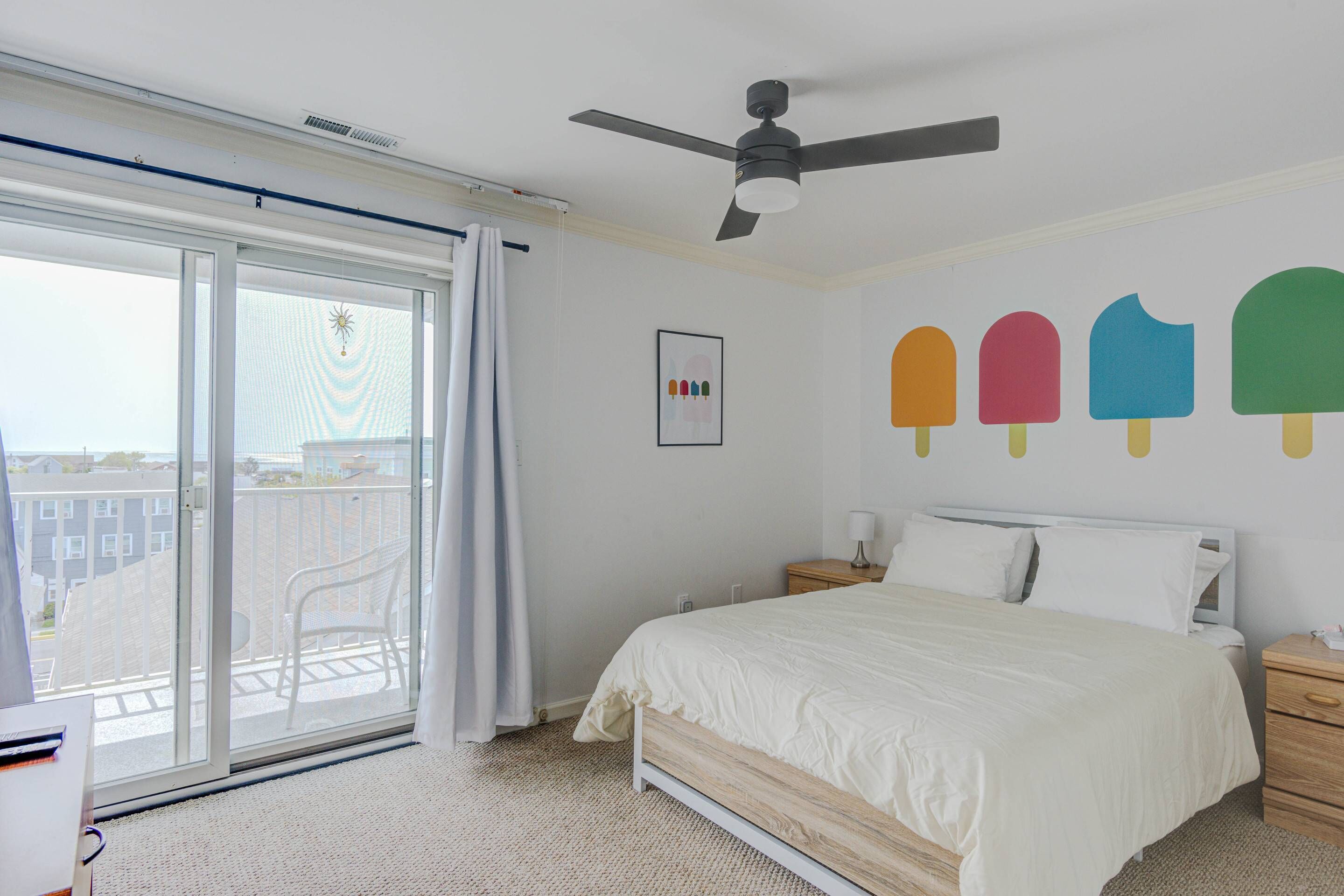 Une chambre avec un lit, un ventilateur de plafond, une fenêtre donnant sur des immeubles et un mur décoré avec des motifs de sucettes glacées.