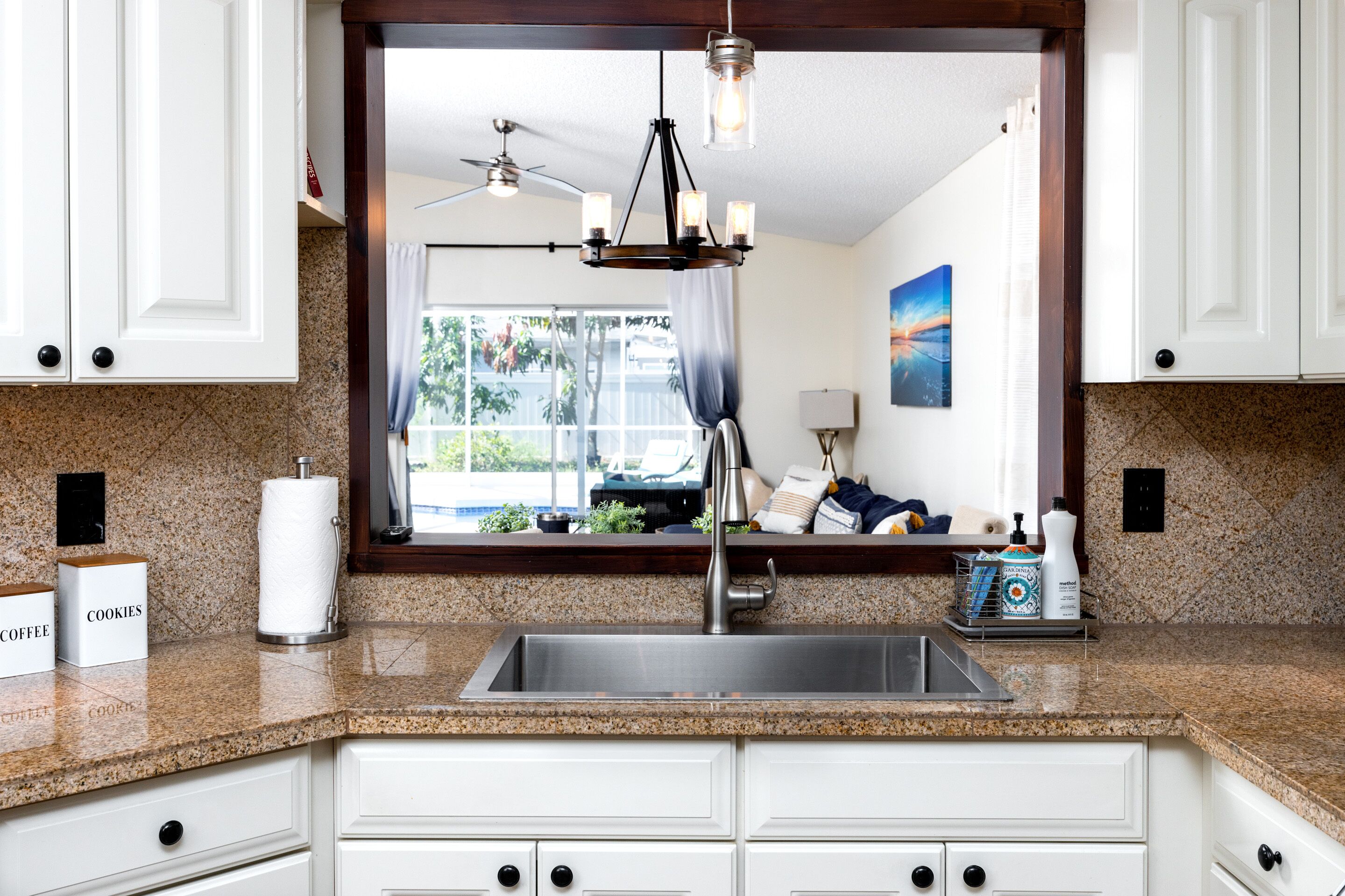 A kitchen with white cabinets, a granite countertop, a stainless steel sink, a chandelier, and a window with a view of the outdoors.