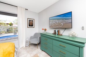 Une pièce comprenant un lit, une commode verte, un fauteuil gris, un tableau encadré, un téléviseur et une fenêtre donnant sur une plage.