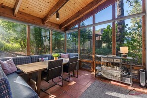 A sunlit room with a wooden ceiling, large windows, a dining table, chairs, a sofa, and a wine rack.