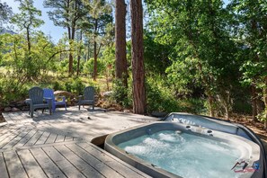 A hot tub surrounded by wooden deck, three Adirondack chairs, and a forested backdrop.