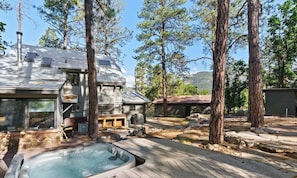 A rustic cabin with a hot tub, surrounded by tall trees and a mountainous backdrop.