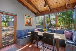 A dining area with a bench, table, and chairs, large windows with a view of trees, and a bear painting on the wall.