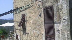 Un edificio de piedra con contraventanas, un pequeño espacio al aire libre con una mesa y sillas, y una sombrilla blanca.