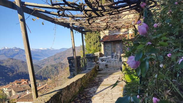 Un sendero de piedra conduce a un edificio de piedra con techo de tejas, rodeado de vegetación y con vistas a un paisaje montañoso.