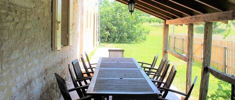 Un patio al aire libre con una larga mesa de madera y sillas, una pérgola arriba y vistas a un jardín y una valla.