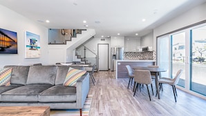 Uma sala de estar moderna com um sofá cinzento, almofadas coloridas, uma mesa de centro de madeira e um tapete riscado. Há uma escada que leva a uma cozinha com uma ilha grande e uma área de jantar com cadeiras.