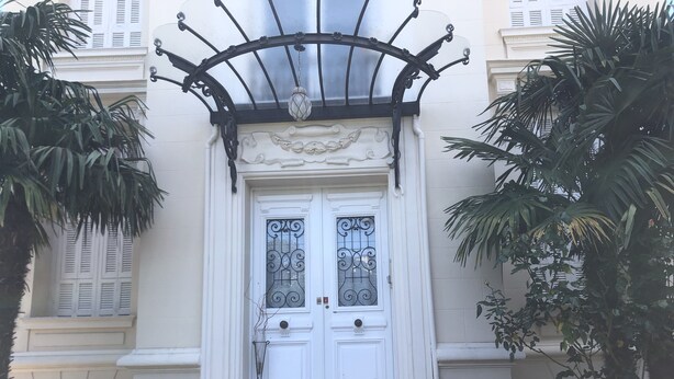 Una porta doble blanca amb detalls metàl·lics decoratius, flanquejada per dues palmeres, que condueix a un conjunt d'escales de marbre.