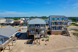 Een strandhuis met meerdere verdiepingen, waaronder een balkon met stoelen, een overdekt terras met tafels en een houten terras.