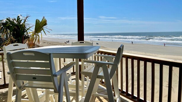 Une terrasse au bord de la plage, avec des chaises blanches et une table, d’où l’on a une vue sur l’océan.