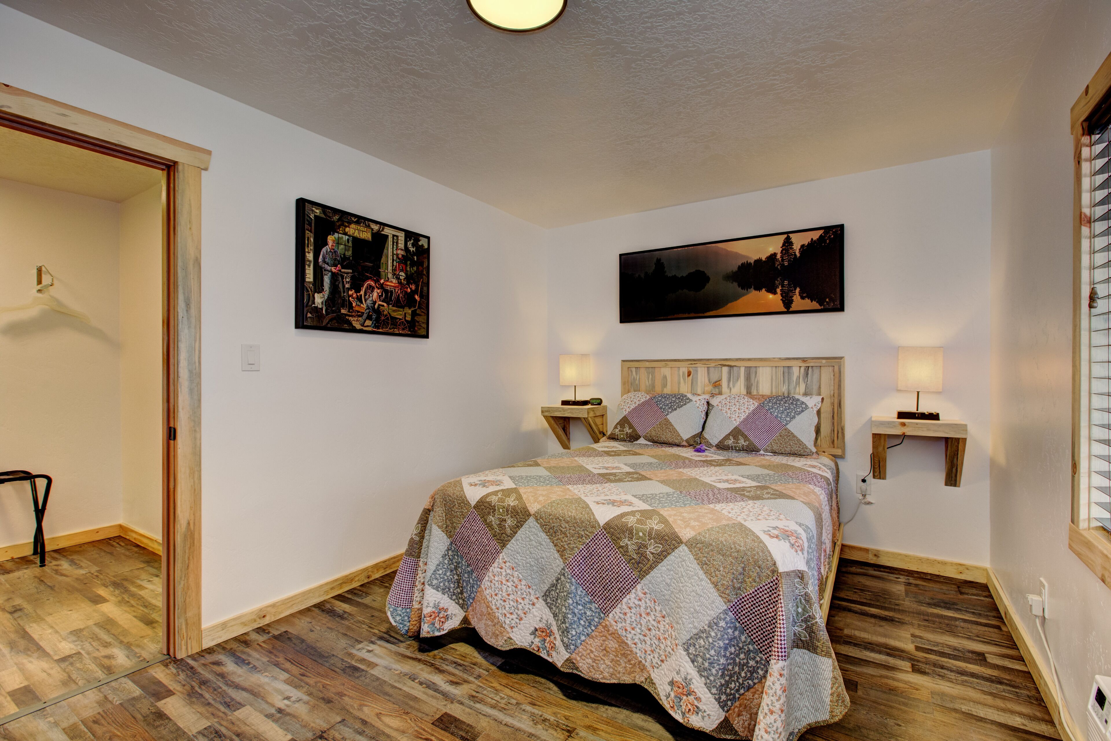 A bedroom with a quilted bedspread, two wall-mounted lamps, and two framed pictures.