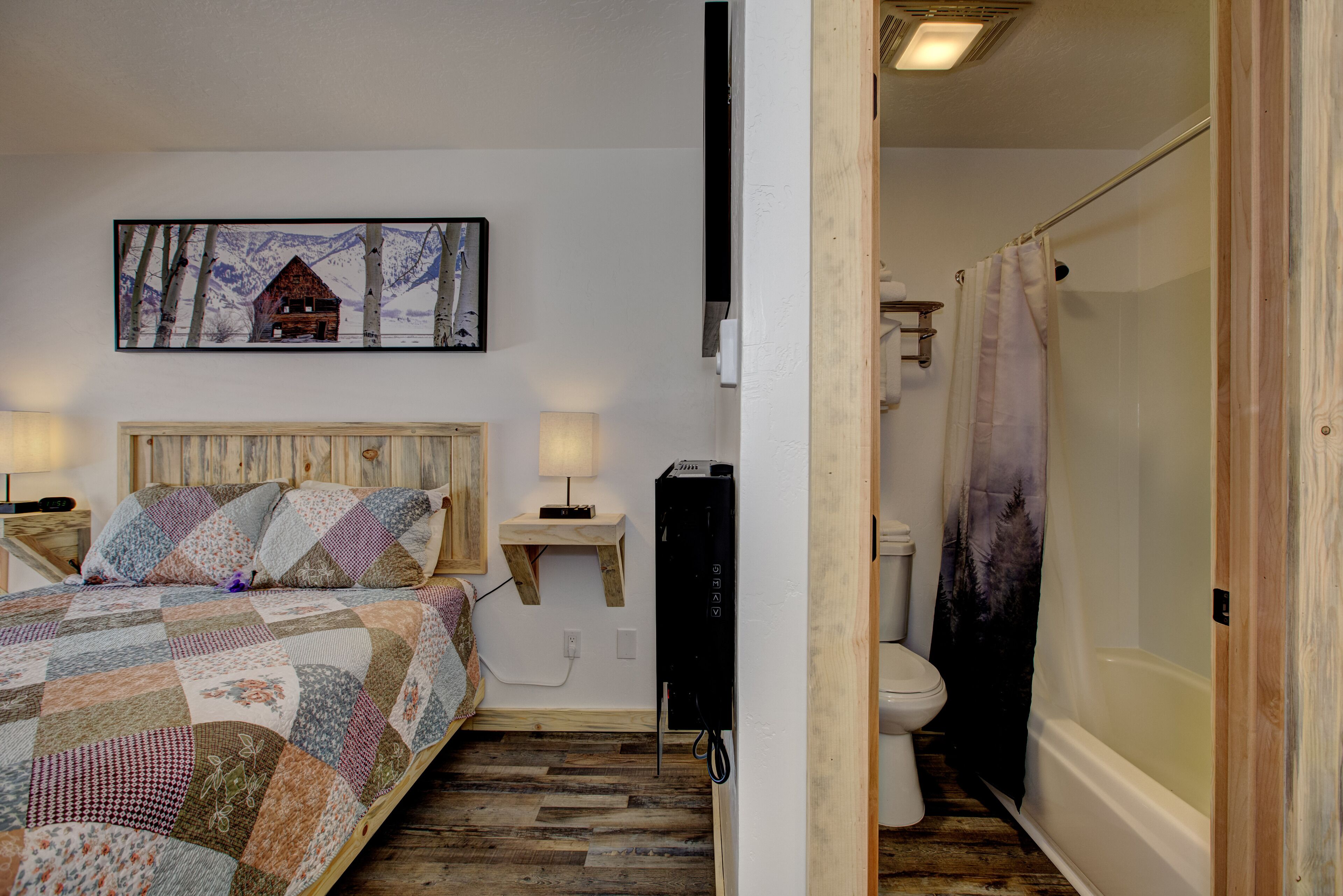 A bedroom with a wooden bed, a mounted TV, a bedside table, and a bathroom with a toilet and bathtub.