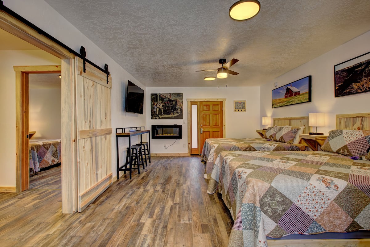 A bedroom with a wooden floor, a bed with a quilt, a desk with a chair, a ceiling fan, and a TV mounted on the wall.