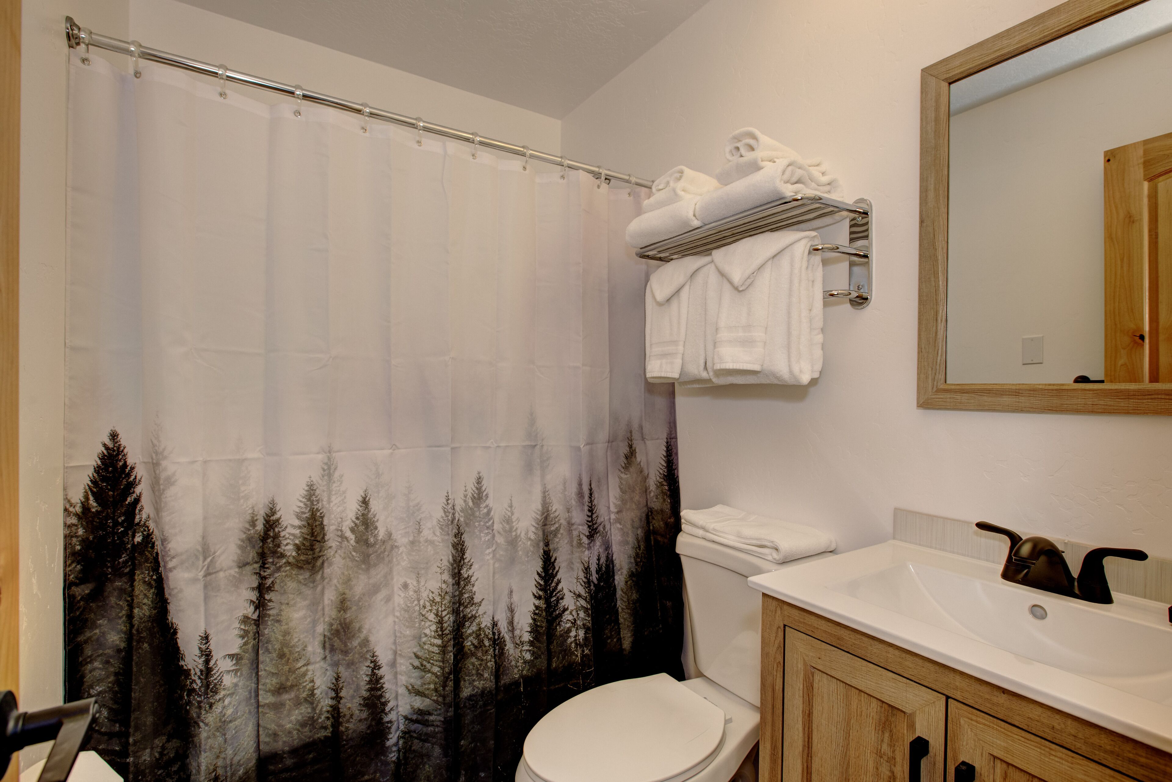 Un baño con cortina de ducha con motivos de un bosque, un inodoro, un lavabo con espejo y un perchero con toallas dobladas.