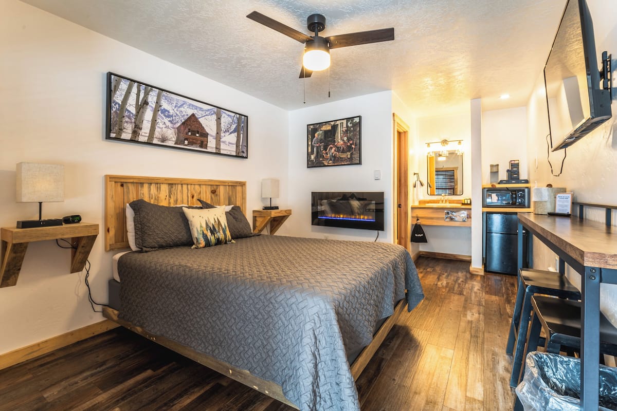 A bedroom with a bed, wooden headboard, wall-mounted TV, ceiling fan, and artwork.