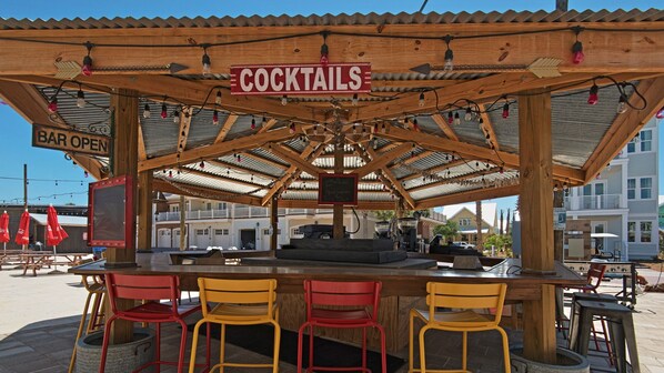 A wooden bar area with a sign reading "COCKTAILS" and a "BAR OPEN" sign. There are four chairs and a counter with a grill.