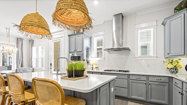 A kitchen with a central island, grey cabinets, wicker chairs, and a hanging light fixture.