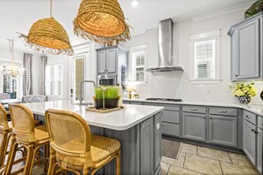 A kitchen with a central island, grey cabinets, wicker chairs, and a hanging light fixture.