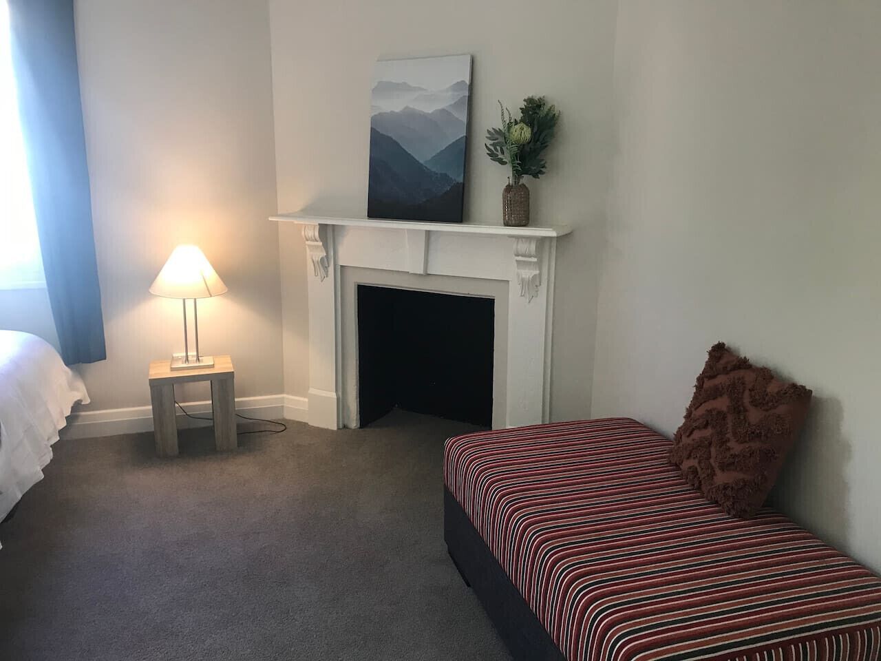 A bedroom with a bed, a nightstand with a lamp, a fireplace, and a painting on the wall.