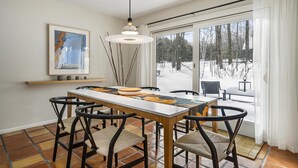 Een eethoek met een tafel, stoelen, een hanglamp, een ingelijst kunstwerk en uitzicht op een besneeerd landschap door een glazen deur.