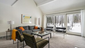 Een moderne woonkamer met een grijze bank, oranje kussens, een glazen salontafel en een groot raam met uitzicht op een besneeerd landschap.